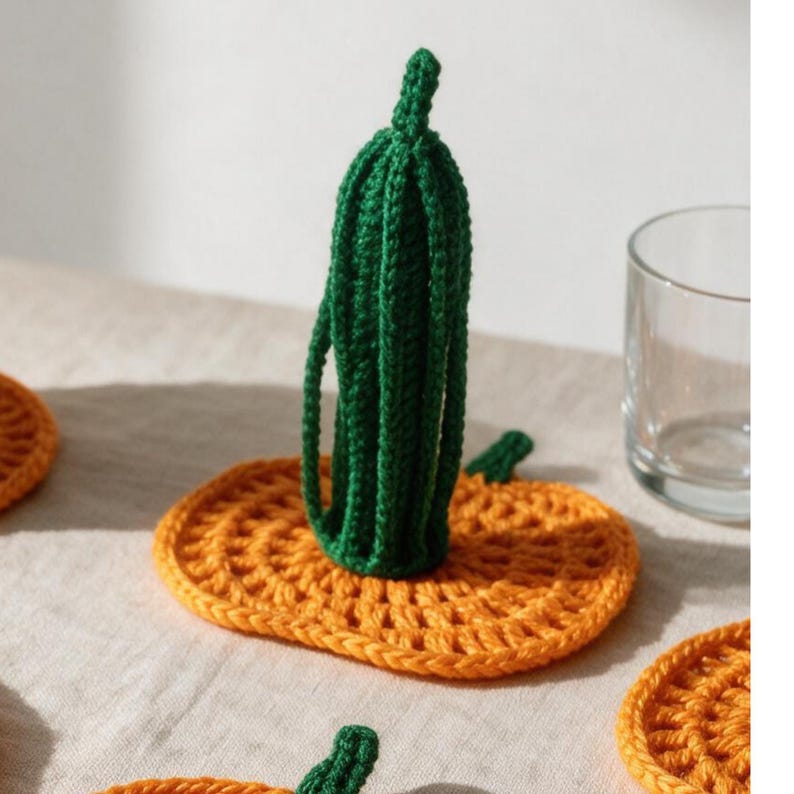 May include: A crocheted orange pumpkin-shaped coaster with a vertical green crocheted stem attached to the center. The coaster features a textured, circular stitch pattern and is crafted from yarn, displaying a vibrant orange base and a dark green stem.