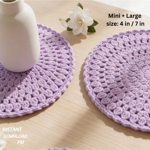 Peut inclure: Deux dessous de plat ronds en crochet lavande, de 10 cm et 18 cm de diamètre, sont présentés sur une surface en bois clair. Un vase blanc et une fleur blanche avec des feuilles vertes sont également présents. Le texte "Mini + Large size: 4 in / 7 in" est visible.