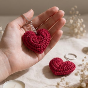 May include: A close-up of two red crocheted heart keychains. One keychain is held in a hand, while the other rests on a surface. The hearts are made of a textured yarn and attached to silver keyrings. The background is softly blurred.