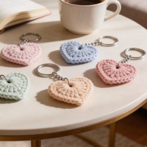 Può includere: Una collezione di portachiavi a forma di cuore in vari colori pastello, tra cui rosa, blu e pesca. Ogni portachiavi è realizzato in materiale all'uncinetto e attaccato a un portachiavi argentato. Una tazza di caffè e un libro sono sullo sfondo.