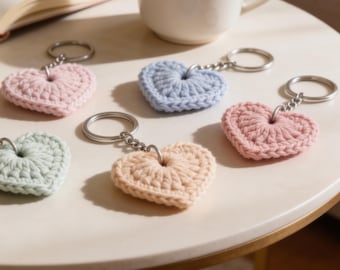 May include: A collection of heart-shaped keychains in various pastel colors, including pink, blue, and peach. Each keychain is crafted from a crocheted material and attached to a silver keyring. A cup of coffee and a book are in the background.