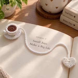 Op de afbeelding: Een witte gehaakte boekenlegger met een miniatuur koffiekopje met een bruin centrum op een schoteltje aan het ene uiteinde en een klein wit gehaakt hartje aan het andere uiteinde, verbonden door een lang wit gehaakt koord op een boekpagina.