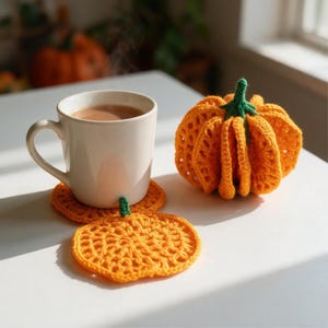 May include: A white ceramic mug filled with a brown beverage sits on a round orange crocheted coaster with a small green stem. Beside it is a three-dimensional orange crocheted pumpkin with a green stem, featuring a textured, open-work pattern.