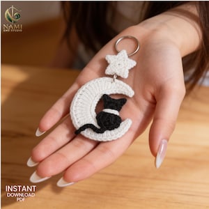 May include: A crochet keyring featuring a white crescent moon and a small white star attached to a silver metal ring. A black crocheted cat is positioned on the centre of the moon. The item is crafted with white and black yarn in a textured stitch.