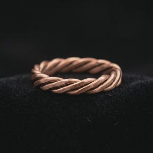 Op de afbeelding: Een koperen ring met een gedraaid, touwachtig ontwerp. Het metaal heeft een matte, getextureerde afwerking met een warme roodbruine tint. De band is opgebouwd uit meerdere strengen die in elkaar zijn gevlochten tot een doorlopend, cirkelvormig vlechtpatroon.