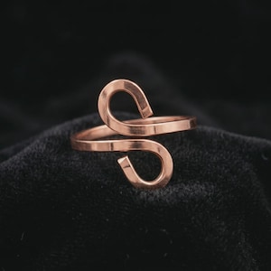 Op de afbeelding: Een open ring in roségoudkleur met een doorlopende band die in het midden buigt in twee tegenovergestelde, afgeronde lussen. Het metaal heeft een gladde, gepolijste afwerking, wat zorgt voor een strak ontwerp dat aan de voorzijde open is.