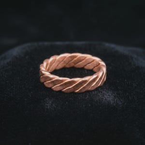 Op de afbeelding: Een koperkleurige ring met een doorlopend gedraaid touwpatroon rondom de gehele band. Het metalen oppervlak heeft een matte, getextureerde afwerking met diagonale groeven die een herhalend gevlochten uiterlijk creëren over de ring.