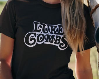 May include: A woman wearing a dark brown t-shirt with the text "LUKE COMBS" in white, smiling next to a white horse. The image is set outdoors with a blue sky and green field in the background.