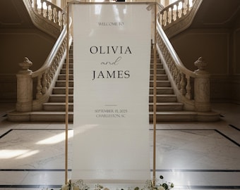 May include: A tall, rectangular white wedding welcome sign held by a wooden frame, featuring the text WELCOME TO OLIVIA and JAMES, SEPTEMBER 15, 2025, CHARLESTON, SC. The base is decorated with a cluster of white roses and green foliage.