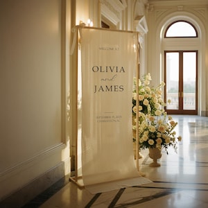 Puede incluir: Un cartel de bienvenida de boda color crema con los nombres Olivia y James, y la fecha 15 de septiembre de 2025, en Charleston, SC. El cartel se muestra en un marco de madera, con arreglos florales cerca.