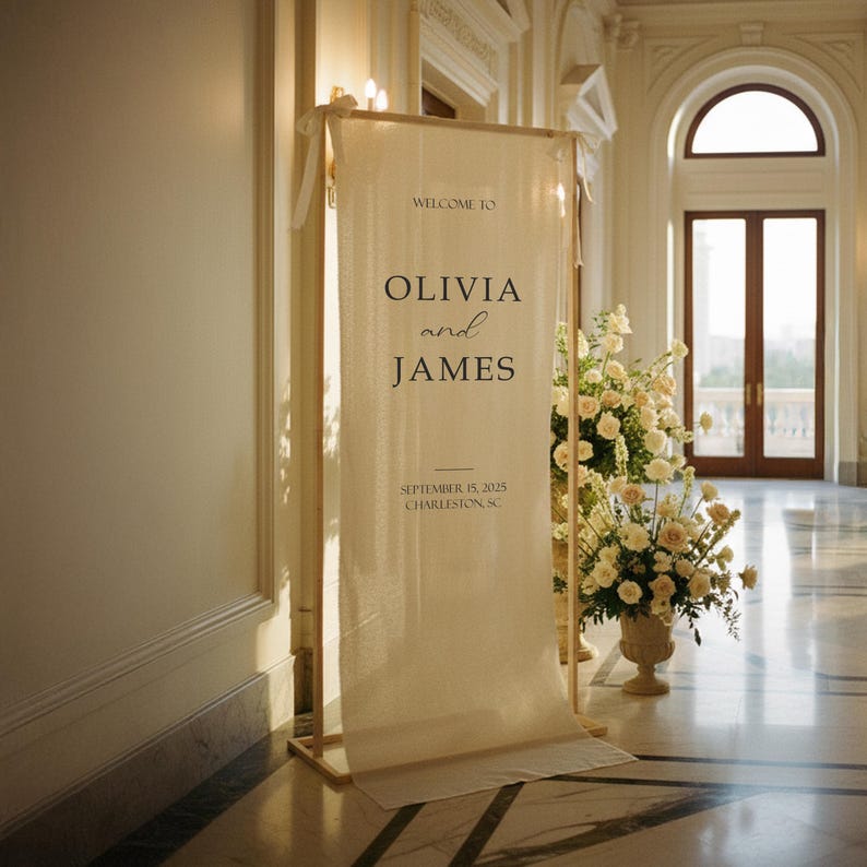 Puede incluir: Un cartel de bienvenida de boda color crema con los nombres Olivia y James, y la fecha 15 de septiembre de 2025, en Charleston, SC. El cartel se muestra en un marco de madera, con arreglos florales cerca.
