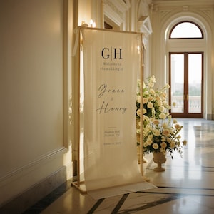 Puede incluir: Un cartel de bienvenida de boda color crema con las iniciales "GH" y los nombres "Grace & Henry". El cartel se muestra en un marco de madera, con arreglos florales y un gran sal&oacute;n al fondo.