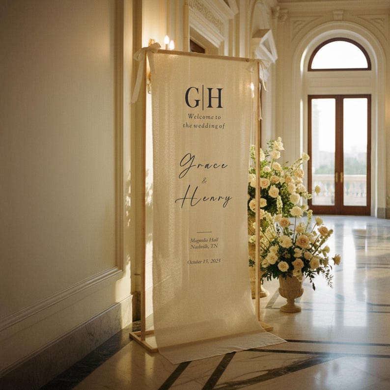 Puede incluir: Un cartel de bienvenida de boda color crema con las iniciales "GH" y los nombres "Grace & Henry". El cartel se muestra en un marco de madera, con arreglos florales y un gran sal&oacute;n al fondo.