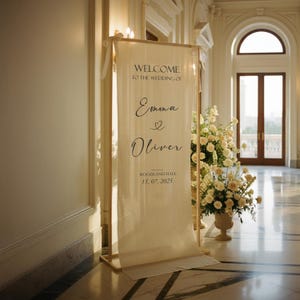 Puede incluir: Un cartel de bienvenida de boda color crema con el texto "WELCOME TO THE WEDDING OF Emma & Oliver" y la fecha "15. 07. 2025". El cartel est&aacute; en un marco de madera, con arreglos florales en el fondo.