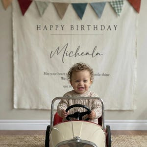 Op de afbeelding: Een crèmekleurige stoffen verjaardagsbanner met de tekst HAPPY BIRTHDAY, de naam Micheala en een citaat, bekroond met een veelkleurige driehoekige vlaggenlijn. Een kind zit in een crèmekleurige en rode metalen speelgoedauto in vintage-stijl.