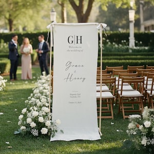 May include: A white wedding welcome sign with the initials "GH" at the top, followed by "Welcome to the wedding of Grace & Henry." The sign is displayed on a wooden frame, with a floral arrangement and chairs in the background.