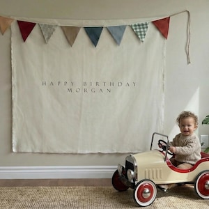 Pode incluir: Uma tapeçaria de tecido cor de creme com o texto HAPPY BIRTHDAY MORGAN em letras pretas, rematada com uma grinalda de bandeirolas triangulares em vermelho, xadrez verde, bege e azul, ao lado de um carro de brinquedo creme estilo vintage.