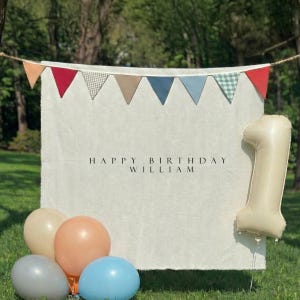 Op de afbeelding: Een rechthoekige gebroken witte stoffen achtergrond met de zwarte tekst HAPPY BIRTHDAY WILLIAM. Boven de achtergrond hangt een slinger van driehoekige vlaggetjes in rood, beige, blauw en ruitpatronen. Een grote crèmekleurige cijferballon één en vier ronde ballonnen.