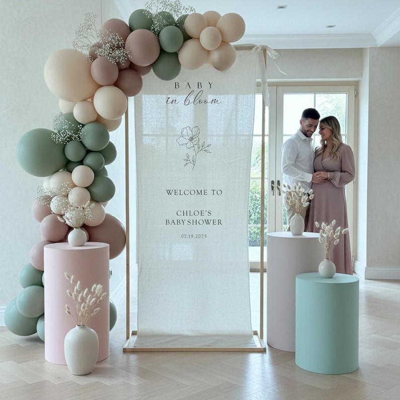 Może przedstawiać: Tablica powitalna na baby shower na drewnianej ramie z napisem BABY in bloom WELCOME TO CHLOE'S BABY SHOWER 07.19.2025, łukiem balonowym w kolorze szałwiowej zieleni, beżu i brudnego r&oacute;żu, cylindrycznymi postumentami oraz suszonymi kwiatami.