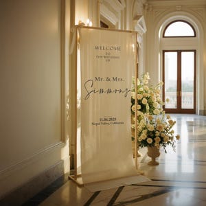 Puede incluir: Un cartel de bienvenida de boda color crema con el texto "WELCOME TO THE WEDDING OF Mr. & Mrs. Simmons" y la fecha 11.06.2025, Nepal Valley, California. El letrero est&aacute; enmarcado en madera clara y est&aacute; en un gran sal&oacute;n.