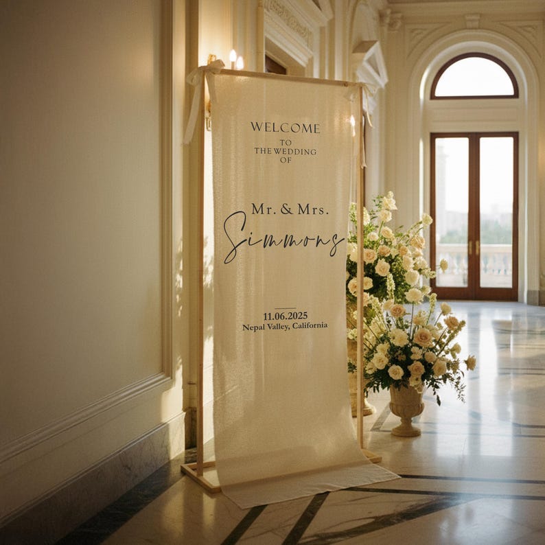 Puede incluir: Un cartel de bienvenida de boda color crema con el texto "WELCOME TO THE WEDDING OF Mr. & Mrs. Simmons" y la fecha 11.06.2025, Nepal Valley, California. El letrero est&aacute; enmarcado en madera clara y est&aacute; en un gran sal&oacute;n.