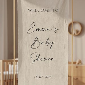 Może przedstawiać: Pionowy baner z beżowej tkaniny zawieszony na drewnianej ramie. Tekst brzmi "WELCOME TO", poniżej "Emma's Baby Shower" pisane kursywą, a na dole data "15. 07. 2025". Tkanina ma widoczny, teksturowany splot materiału.