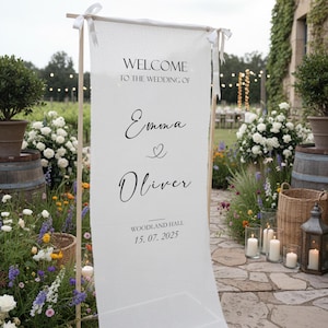 Puede incluir: Un letrero de boda de tela blanca con el texto "WELCOME TO THE WEDDING OF Emma & Oliver" en escritura negra. El letrero est&aacute; sostenido por postes de madera y atado con cintas blancas. Tambi&eacute;n se muestra la fecha "15. 07. 2025". El letrero est&aacute; instalado al aire libre.