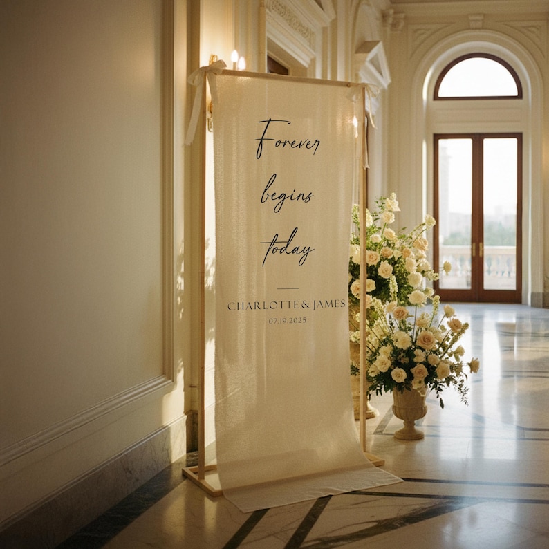 Puede incluir: Una pancarta de boda color crema con el texto "Forever begins today" en escritura elegante. Debajo, se lee "CHARLOTTE & JAMES" y la fecha "07.19.2025". La pancarta se muestra en un marco de madera.
