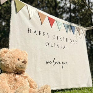 May include: A beige fabric banner featuring a colourful pennant flag border at the top. The text reads HAPPY BIRTHDAY OLIVIA! and we love you in black lettering. A light brown plush teddy bear with a satin ribbon bow sits in the foreground.