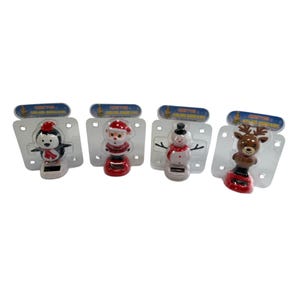 May include: Four solar-powered Christmas ornaments: a penguin, Santa Claus, a snowman, and a reindeer. Each figure is mounted on a red base and packaged on a clear plastic card with a blue header. The penguin has a red hat and scarf.