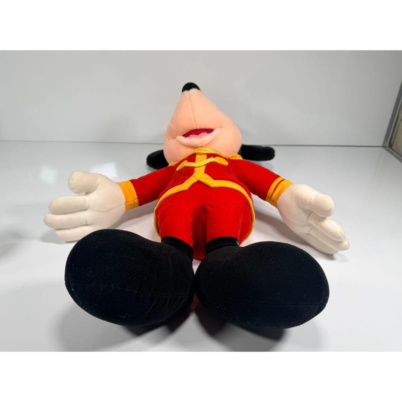 May include: A plush Mickey Mouse doll in a red and yellow outfit with black shoes and ears. The doll has white gloves and is lying on its back with its arms outstretched. The doll's face is a light pink color.