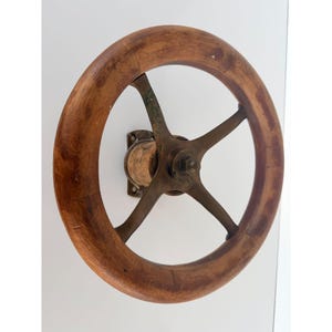 May include: A circular handwheel featuring a polished wooden rim and a central brass mechanism with four spokes. The assembly is mounted to a square brass base plate with four screw holes, showcasing a metallic finish and natural wood grain texture.