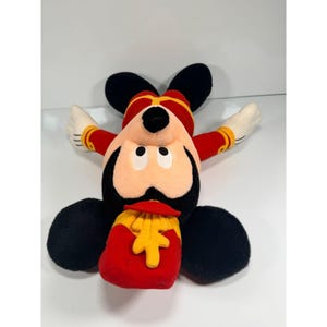 May include: A plush Mickey Mouse toy in a red and gold band uniform. The toy has black ears, a black face, and a red and gold hat. Mickey is holding a red bag with yellow fries.