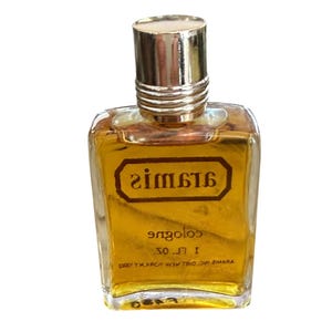 May include: A vintage cologne bottle with a gold-colored liquid inside. The bottle has a gold-colored cap and a label with the brand name "Aramis" in a brown rectangular frame. The bottle is 1 fl oz.