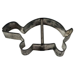 Op de afbeelding: Een vintage metalen koekjessnijder in de vorm van een schildpad. De snijder heeft een eenvoudige omtrek met een kop, vier poten en een staart. Het metaal heeft een versleten, verouderd uiterlijk.