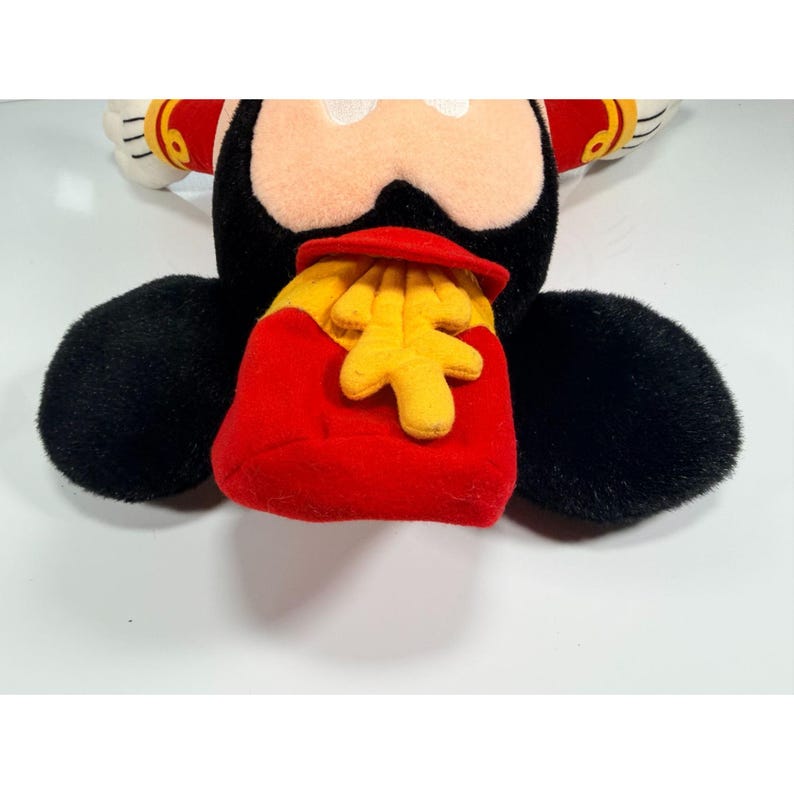 May include: A plush Mickey Mouse toy with black fur, a red and yellow outfit, and a red bag of yellow fries. The toy has white gloves and a smiling face. The toy is on a white surface.