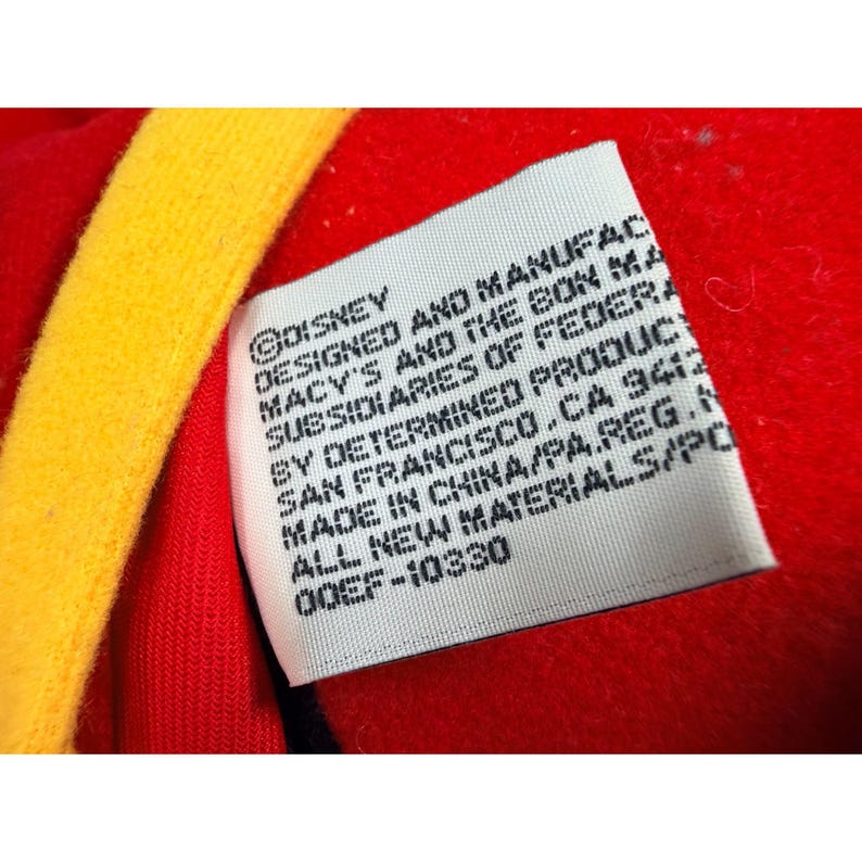 May include: Close-up of a tag with text. The tag is white with black text and is attached to a red fabric. The text includes "&copy;DISNEY", "DESIGNED AND MANUFAC", and "MADE IN CHINA/PA.REG.N". A yellow fabric is visible.