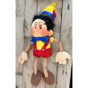 May include: A vintage Pinocchio doll with a yellow shirt, red shorts, and a blue bow tie. The doll has a yellow hat with blue and red accents, black hair, and wooden arms and legs. The doll's hands and feet are made of a cream-colored fabric.
