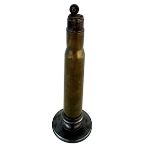May include: A vintage brass lighter fashioned from a repurposed metallic cartridge casing. The cylindrical body features a weathered patina, topped with a dark metal ignition mechanism and a circular base for stability on a flat surface.