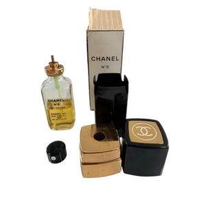 May include: A collection of Chanel No. 5 perfume items. Includes a perfume bottle, a black cap, a gold-coloured perfume holder, a black and gold compact, and a box. The bottle contains a yellow liquid and the text "CHANEL N&deg;5".