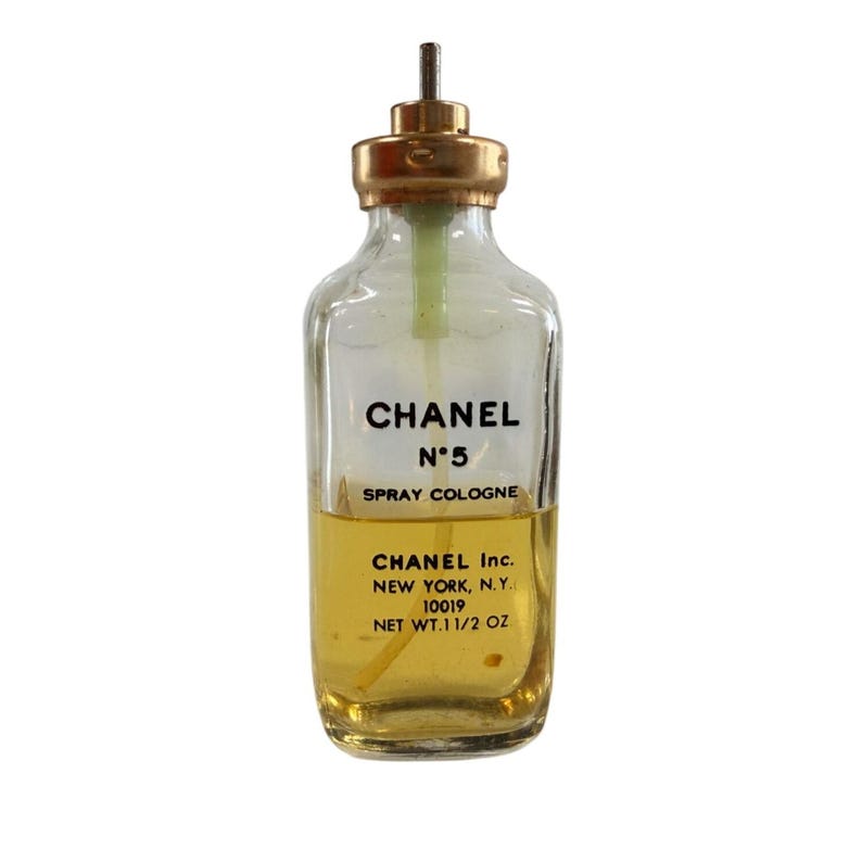 May include: A vintage Chanel No. 5 spray cologne bottle. The clear glass bottle has a gold-coloured cap and contains a yellow liquid. The label reads "CHANEL N&deg;5 SPRAY COLOGNE" and "CHANEL Inc. NEW YORK, N.Y. NET WT.1 1/2 OZ."