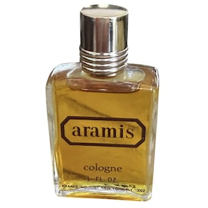 May include: A small, square cologne bottle with a silver cap and a brown label. The label reads "aramis cologne 1 FL. OZ." The bottle contains a golden-colored liquid. The bottom of the bottle has text that reads "ARAMIS INC., DIST NEW YORK, NY 10022."