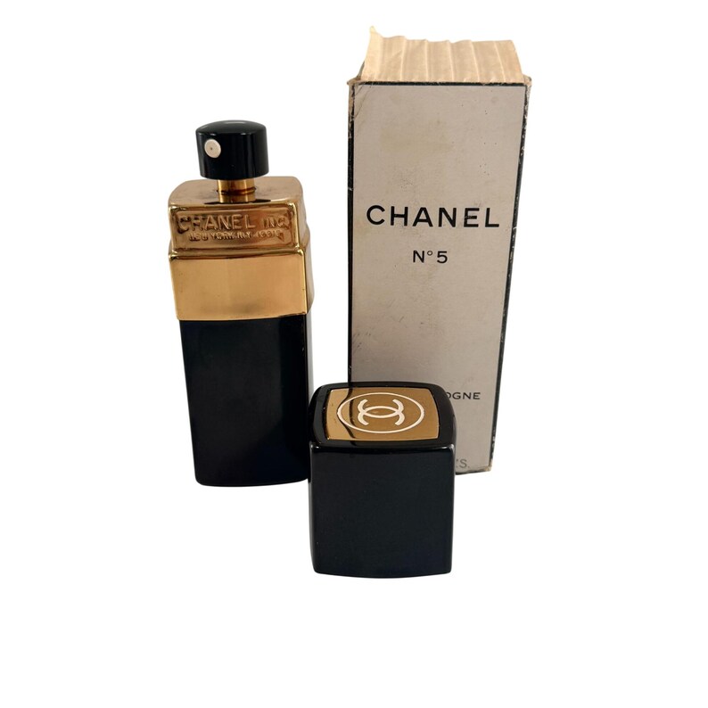 May include: A black and gold Chanel No. 5 perfume bottle, cap, and box. The bottle has a gold top and a black base. The box is white with black text that reads "CHANEL N&deg;5 COLOGNE". The cap is black with a gold emblem.