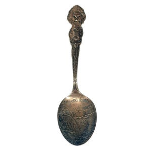 May include: A silver spoon with a detailed design. The handle features a Native American figure at the top and a detailed pattern. The spoon's bowl depicts Niagara Falls with the words "Niagara Falls" above the image.