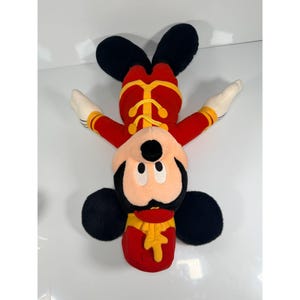 May include: A plush Mickey Mouse toy in a red and yellow band uniform. The toy has black ears, a black face, and white gloves. The toy is lying upside down on a white surface.
