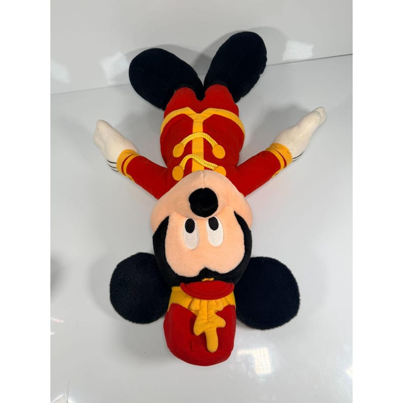May include: A plush Mickey Mouse toy in a red and yellow band uniform. The toy has black ears, a black face, and white gloves. The toy is lying upside down on a white surface.