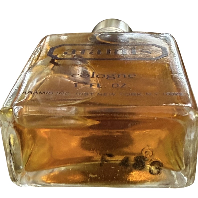 May include: A vintage cologne bottle with a clear, geometric design. The bottle contains a golden-brown liquid and features the brand name "Aramis" in a rectangular label. The label also indicates the contents are 1.7 fl oz.