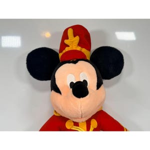 May include: A plush Mickey Mouse toy wearing a red and yellow band uniform. The toy has black ears, a peach-colored face, and a red hat with yellow accents. The uniform has gold trim.