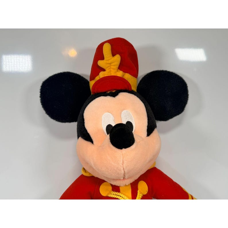 May include: A plush Mickey Mouse toy wearing a red and yellow band uniform. The toy has black ears, a peach-colored face, and a red hat with yellow accents. The uniform has gold trim.