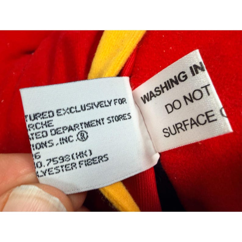 May include: Close-up of two white fabric tags with black text against a red and yellow background. One tag reads "WASHING IN DO NOT SURFACE". The other tag has text that reads "TURED EXCLUSIVELY FOR RCKE TED DEPARTMENT STORES MONS.INC. 0.7598(HK) LYEESTER FIBERS".